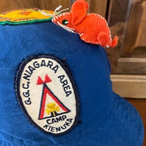 May include: A blue baseball cap featuring a patch with "G.C. Niagara Area Camp Kienuka". An orange felt mouse is attached to the cap. The cap also has a yellow and green patch that reads "Camp Niagara".