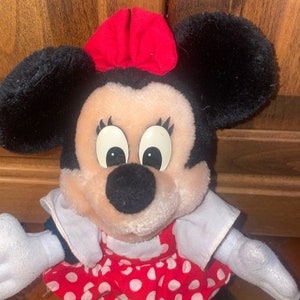May include: A plush Minnie Mouse toy wearing a red bow and a red and white polka dot dress.