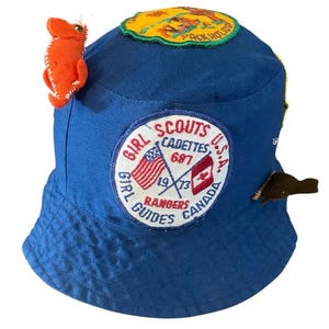 Puede incluir: Un sombrero de cubo azul de las Girl Scouts con parches. El sombrero presenta un caballito de mar de fieltro naranja cosido, un parche circular con las palabras "Girl Scouts Cadettes U.S.A. Rangers Canada" y un parche amarillo y verde.