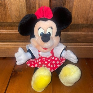 May include: A plush Minnie Mouse toy wearing a red dress with white polka dots and a red bow. The toy has black ears, a black nose, and yellow shoes.