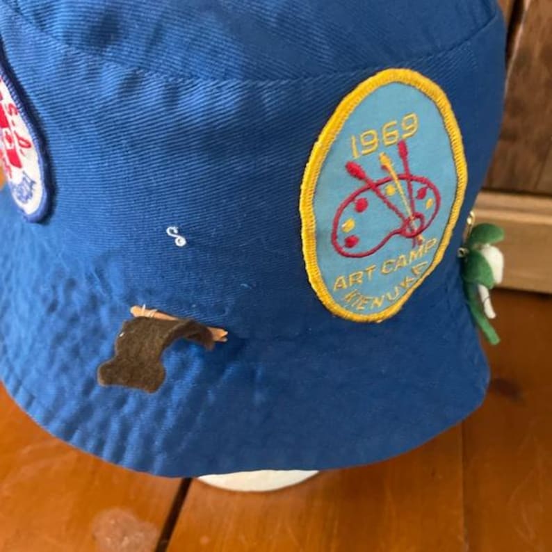 May include: Blue bucket hat with embroidered patches. One patch reads "1969 ART CAMP MENU". The hat has a small brown patch and a green and white charm attached. The hat is made of a soft, durable fabric.