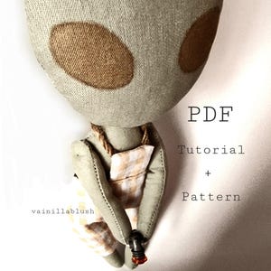 May include: A PDF pattern and tutorial for sewing a gray alien doll with brown eyes, wearing a yellow and white checkered dress. The doll is holding a small orange bead.