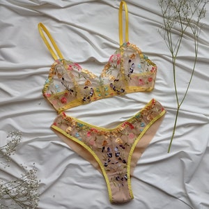 May include: A yellow and nude lingerie set with floral embroidery. The bra has adjustable straps and a triangle cup shape. The matching panty has a high-cut leg and a low rise.