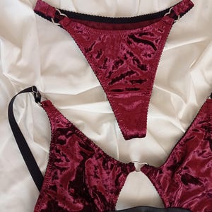 May include: A burgundy velvet lingerie set with black straps and silver heart-shaped rings. The set includes a bra and a thong.