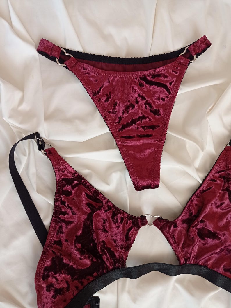 May include: A burgundy velvet lingerie set with black straps and silver heart-shaped rings. The set includes a bra and a thong.