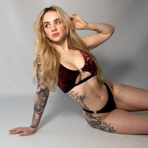 May include: A woman wearing a burgundy velvet lingerie set with black trim and a heart-shaped ring detail. She has tattoos on her arms and torso.