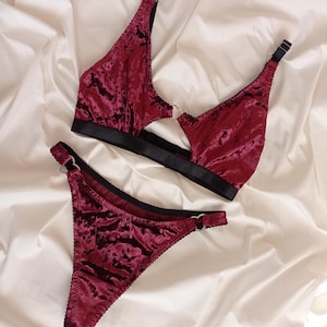 May include: A burgundy velvet lingerie set with black trim and silver rings. The set includes a bra and a thong.