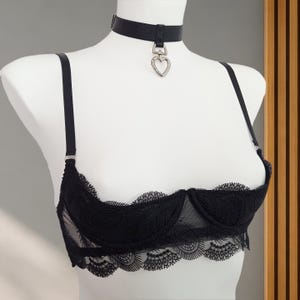 Può includere: Un reggiseno nero in pizzo a coppe aperte con sottili spalline regolabili e un girocollo nero abbinato con un ciondolo argentato a forma di cuore. Il reggiseno presenta ferretti di sostegno e dettagli in pizzo smerlato sul bordo inferiore.