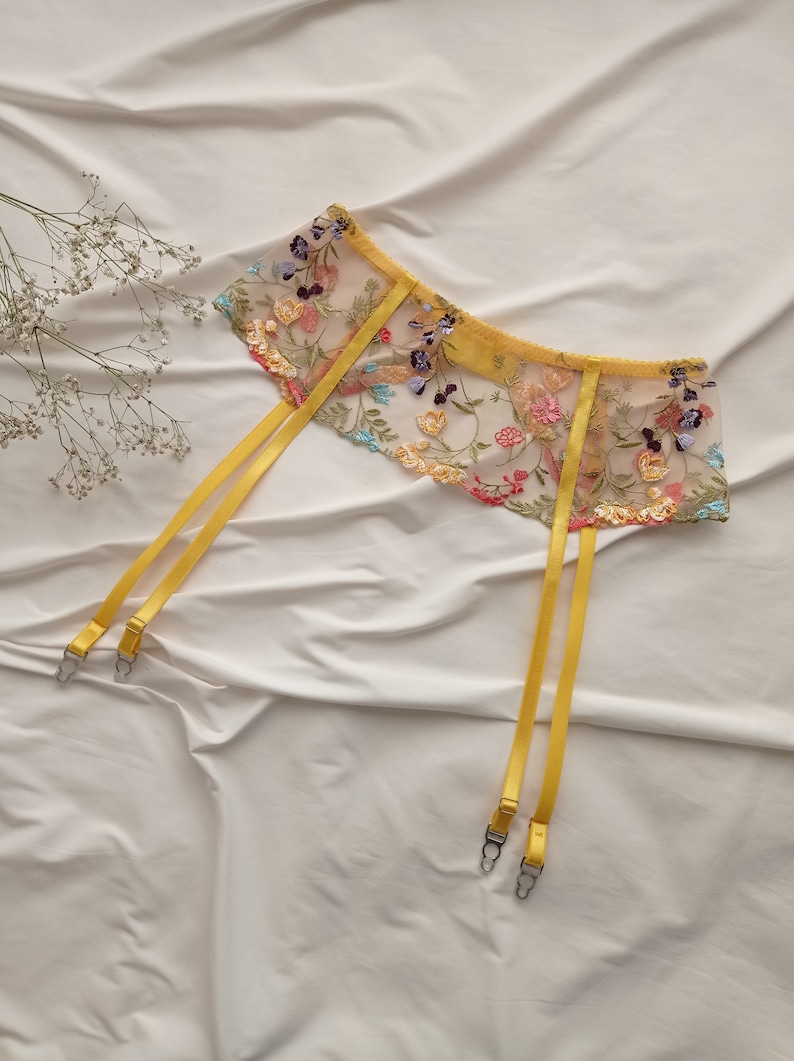 May include: Yellow floral embroidered suspender belt with adjustable straps. The suspender belt is made of delicate lace and features a floral pattern in shades of pink, blue, and green.