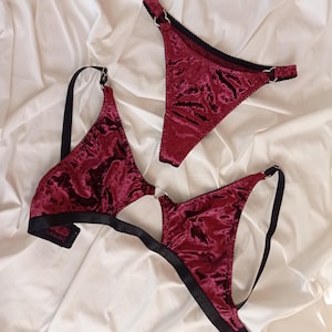 May include: A burgundy velvet lingerie set with black trim and silver rings. The set includes a bra and a thong.