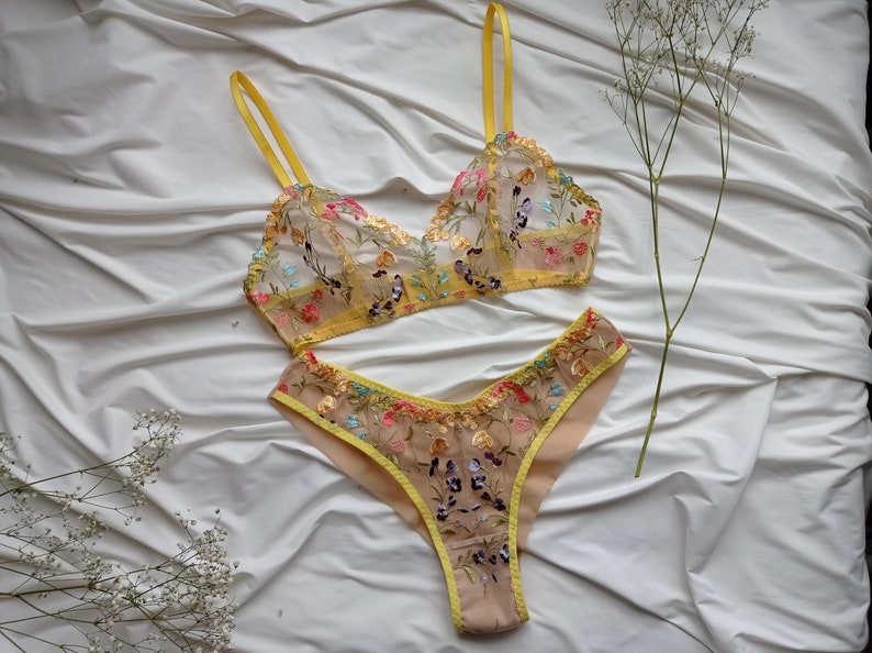 May include: A yellow and nude lingerie set with floral embroidery. The bra has adjustable straps and a triangle cup shape. The matching panty has a high-cut leg and a low rise.