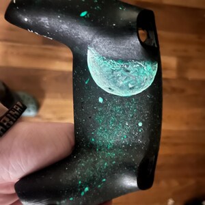 May include: Black controller with a space-themed design. The controller features a painted galaxy design with a teal-colored planet and green speckles. The controller is held in a hand.