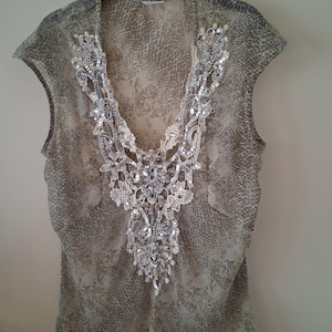 Peut inclure: Un chemisier sans manches avec un col en V, orné d'un motif peau de serpent beige et gris. Le décolleté est orné de sequins argentés et de dentelle, ajoutant une touche décorative. Le chemisier est suspendu à un cintre noir.
