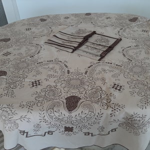 Op de afbeelding: Een ronde, witte tafelkleed met een bruin bloemenpatroon. Het tafelkleed heeft een gegolfde rand en is versierd met een set bruine servetten.