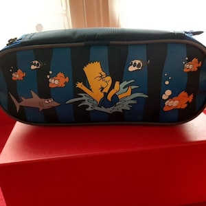 May include: A blue pencil case with a cartoon image of a yellow character swimming with fish and sharks. The case is on a red box.