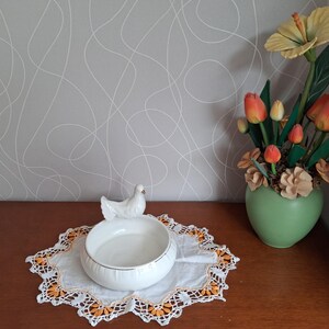 Puede incluir: Un cuenco de cerámica blanco con una figura de paloma encima, sobre un mantelito blanco con ribetes naranjas y amarillos. A la derecha, un jarrón con tulipanes naranjas y una flor amarilla.