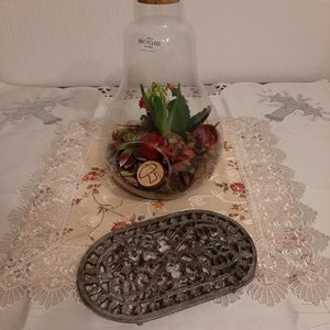 Puede incluir: Una botella de vidrio transparente con un tapón de corcho, llena de artículos decorativos, incluyendo una pequeña planta, bayas rojas y setas de madera. La botella está sentada sobre un salvamanteles de metal gris con un diseño ornamentado. La botella tiene una etiqueta que dice "100% Vidrio Reciclado".