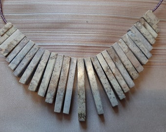 May include: A necklace made of light brown, rectangular, stone-like beads. The beads are arranged in a fan shape, with the longest beads at the bottom and the shortest beads at the top. The necklace is made of a light brown, rectangular, stone-like beads.