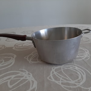 Könnte beinhalten: Ein silberner Topf mit einem braunen Griff. Der Topf steht auf einem Tisch mit einer weißen und beigefarbenen Tischdecke.
