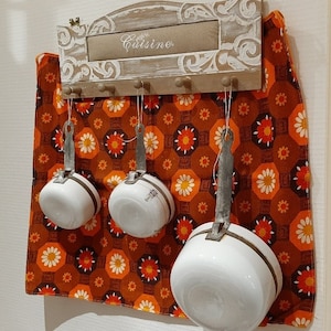 May include: A white wooden rack with the word "Cuisine" written on it hangs on a wall. The rack has hooks for hanging kitchen utensils. Three white enamel pots with metal handles are hanging from the hooks. The wall behind the rack is covered in orange and brown patterned fabric.