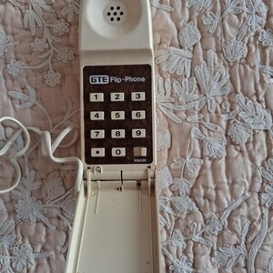 May include: An off-white GTE Flip-Phone with a flip-down keypad. The phone has a series of numbered buttons and the word "RINGER" is printed on the bottom. The phone is connected to a white cord.