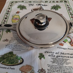 Könnte beinhalten: Ein silberner Topf aus Edelstahl mit einem schwarzen Griff und einem Glasdeckel. Der Topf steht auf einem weißen Küchentuch mit grünem und weißem Muster. Das Tuch hat ein Rezept für Pesto alla genovese darauf gedruckt.