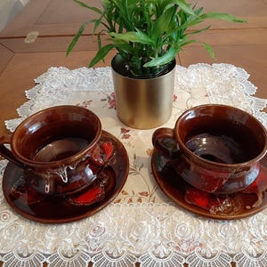 Può includere: Due tazze in ceramica marrone con smalto rosso e marrone, ciascuna su un piattino abbinato, su un tovaglia di pizzo bianco.