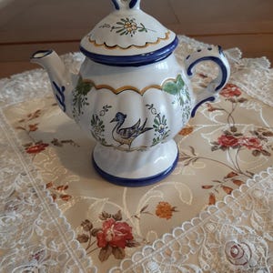 Op de afbeelding: Een witte theepot van keramiek met een blauwe en gouden rand. De theepot heeft een bloemenmotief en een vogel die aan de zijkant is geschilderd. De theepot staat op een wit kantkleed.