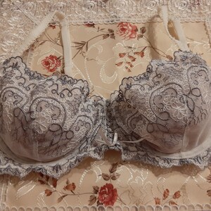 May include: A delicate lace bra with a white base and gray embroidery. The bra has a scalloped edge and a bow in the center.