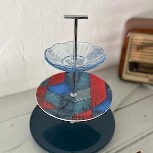 May include: A three-tiered serving stand with a chrome handle. The top tier is a scalloped, light blue glass plate. The middle tier has a geometric pattern in red, blue, and gray. The bottom tier is a solid, dark teal plate.