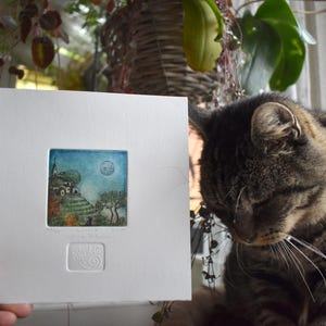 May include: A small, framed print featuring a colorful landscape with a church, trees, and a full moon. The print is held in front of a tabby cat. The artwork is in a white mat with a small embossed design.