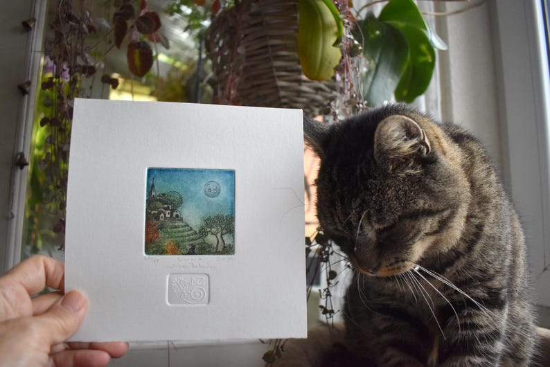 May include: A small, framed print featuring a colorful landscape with a church, trees, and a full moon. The print is held in front of a tabby cat. The artwork is in a white mat with a small embossed design.