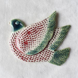 Può includere: Un ornamento di colomba in ceramica con ali macchiate di rosso e bianco e dettagli di foglie verdi. La colomba ha un occhio blu e bianco.