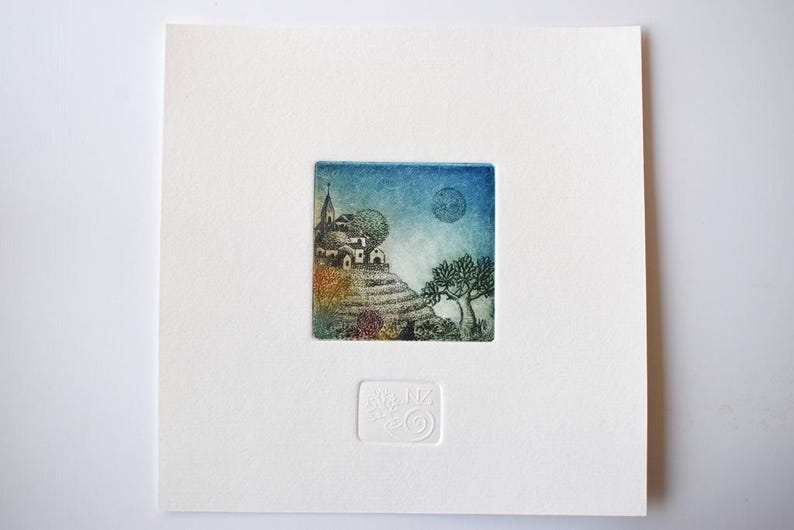May include: A small, square art print featuring a whimsical landscape with a village on a hill, trees, and a moon in a blue sky. The print is set on a white background with a textured, embossed design.
