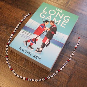 以下が含まれることがあります： レイチェル・リード著「The Long Game」というタイトルの本。表紙には2人のホッケー選手が描かれています。「RTMH ATTSSTINE」と「ATTS SSS RTMH」と書かれたビーズネックレスは、白い文字ビーズ、赤と青のビーズで構成されています。