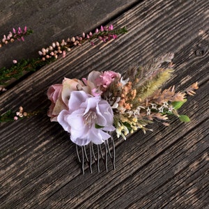 May include: A floral hair comb with a pink and white flower, dried grasses, and small white flowers. The comb is on a rustic wooden background.