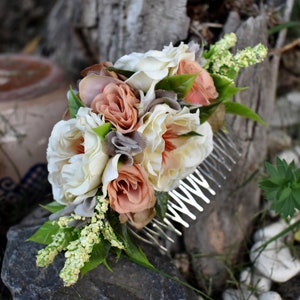 Puede incluir: Un peine para el cabello floral con flores de tela blancas y melocotón. El peine tiene una base de metal plateado con dientes.