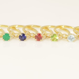 May include: A collection of gold-colored rings, each featuring a different colored gemstone. The gemstones include turquoise, pearl, ruby, pink, emerald, sapphire, red, peridot, aquamarine, opal, diamond, and amethyst. The rings are displayed on a white surface.