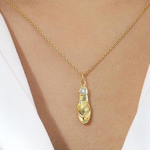 May include: A gold-toned necklace featuring a delicate chain and a pendant shaped like a single flip-flop sandal. The pendant is accented with a small, clear, sparkling stone at the top, reflecting light against the polished metallic surface.