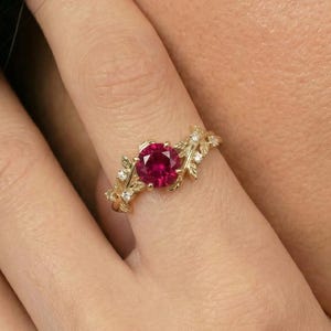 May include: A gold ring featuring a large, round, red gemstone at the center, surrounded by smaller white gemstones and leaf-like designs. The ring is on a finger.