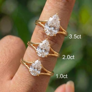 Op de afbeelding: Drie gouden ringen met peervormige diamanten van verschillende maten worden op een vinger getoond. De ringen zijn gelabeld met hun karaatgewichten: 3,5 ct, 2,0 ct en 1,0 ct. De achtergrond is een wazig groen.