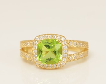 May include: A gold ring featuring a square, light green gemstone at the center, surrounded by smaller, clear gemstones. The band splits into two rows of clear gemstones on each side. A jewelry piece.