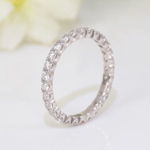 May include: A silver eternity ring featuring a continuous line of small, round, clear gemstones. The ring is set against a soft, white background, with a blurred white flower in the background.