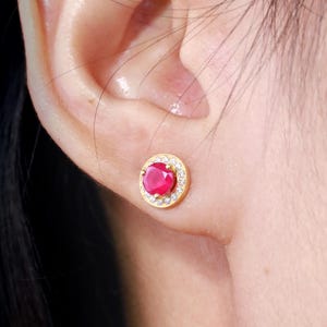 Może przedstawiać: Para złotych kolczyków z czerwonym kamieniem otoczonym halo mniejszych białych kamieni.