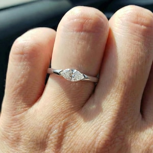 May include: A silver ring with a marquise-cut diamond. The diamond is set horizontally on a thin band. The ring is on a finger, with the background out of focus.