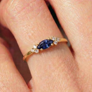 May include: A gold ring featuring a marquise-cut, deep blue gemstone as the centerpiece, flanked by smaller, round, clear gemstones. The ring is set on a delicate gold band, showcasing a classic and elegant design.