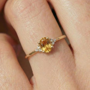 Op de afbeelding: Een gouden ring met een ovale, gele edelsteen als middelpunt. Kleine, heldere accentstenen zijn aan weerszijden van de hoofdsteen gezet. De ring is gezet op een delicate gouden band, met een klassiek en elegant ontwerp.