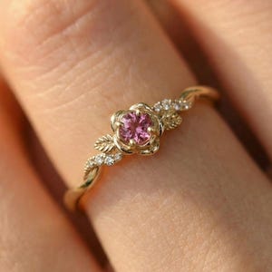 May include: A gold-colored ring featuring a pink gemstone flower at the center. The ring has a delicate, twisted band with small, clear accent stones and leaf details on either side of the flower.