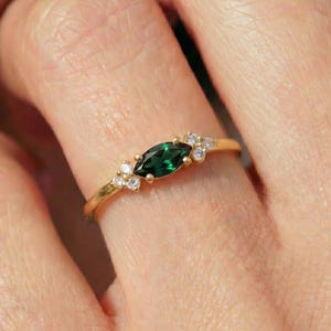 May include: A gold ring featuring a marquise-cut emerald green gemstone, flanked by small, round, clear gemstones. The ring is set on a delicate gold band, showcasing a classic and elegant design.