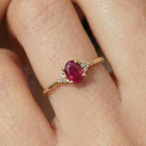 May include: A gold ring featuring an oval-cut ruby gemstone. The ruby is a deep red color and is flanked by small, clear accent stones. The ring's band is a thin, polished gold.